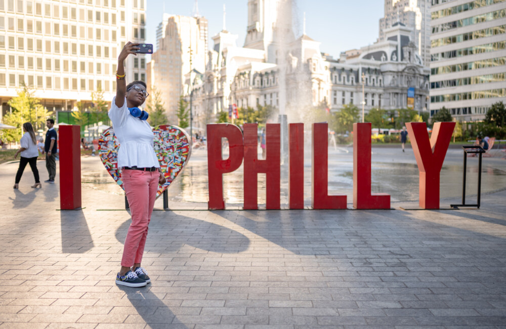 A person takes a selfie in front of a 3D sculpture spelling the message "I 'heart' Philly."