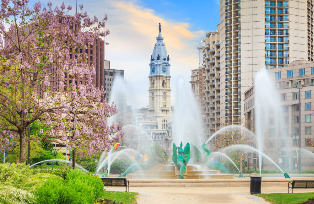 Active fountain in a paved plaza with a tree in full bloom on the left and tall buildings in the background.