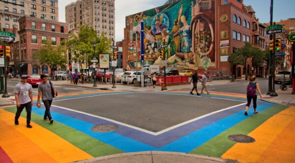People walk across rainbow-colored crosswalks installed in a neighborhood filled with large brick buildings, one featuring a large and colorful mural.