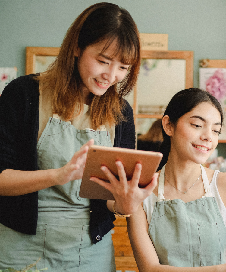 Two women wearing aprons looking at computer and ipad in small business..