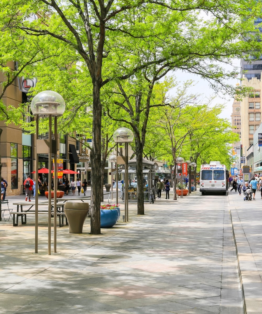 The 16th street mall in downtown with shops and cafes and green trees in the middle of the street. People are doing shopping, walking by or dining.