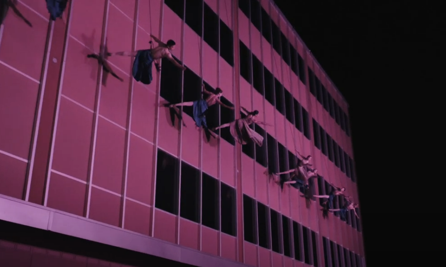 Dancers move suspended from a wall bathed in pink light