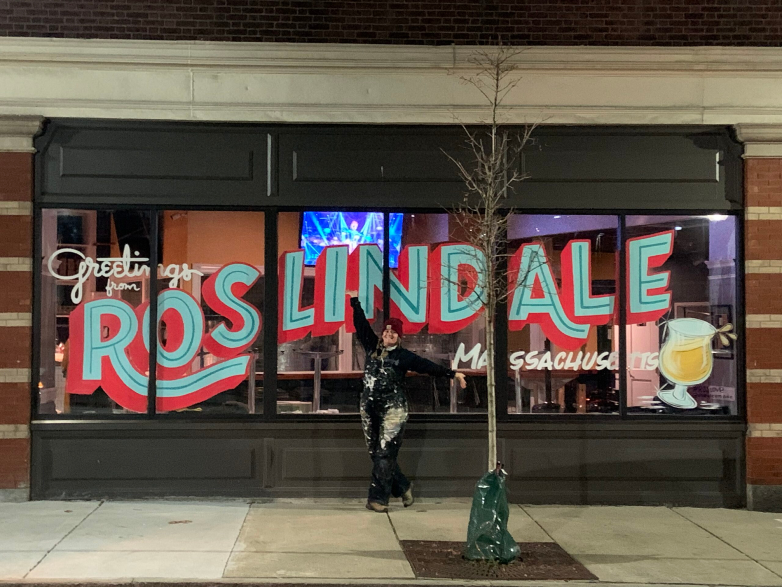 A woman poses in front of a window mural reading "Greetings from Roslindale Massachusetts"