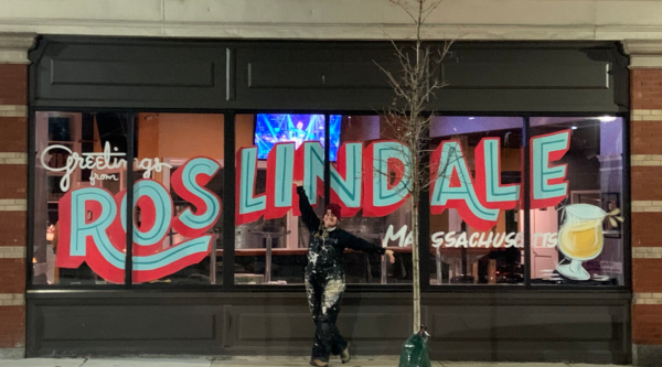 A woman poses in front of a window mural reading "Greetings from Roslindale Massachusetts"