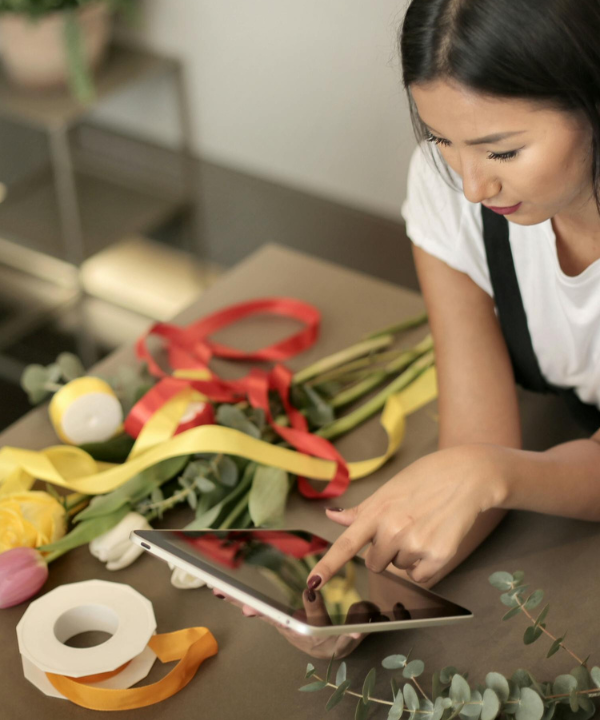 Woman with black hair wearing apron and leaning on work bench covered with floral arrangement supplies looks at ipad.