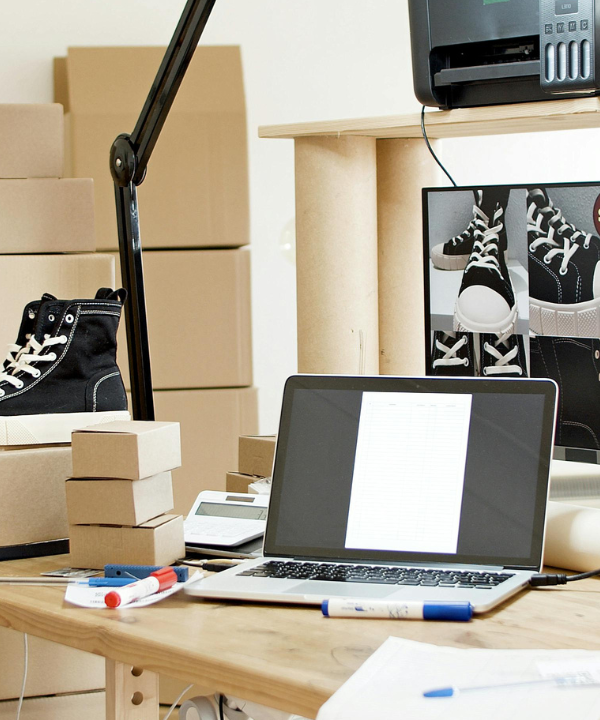 Boxes sitting on desk with sneaker on top of one box and computer screens showing mock-up of sneaker.