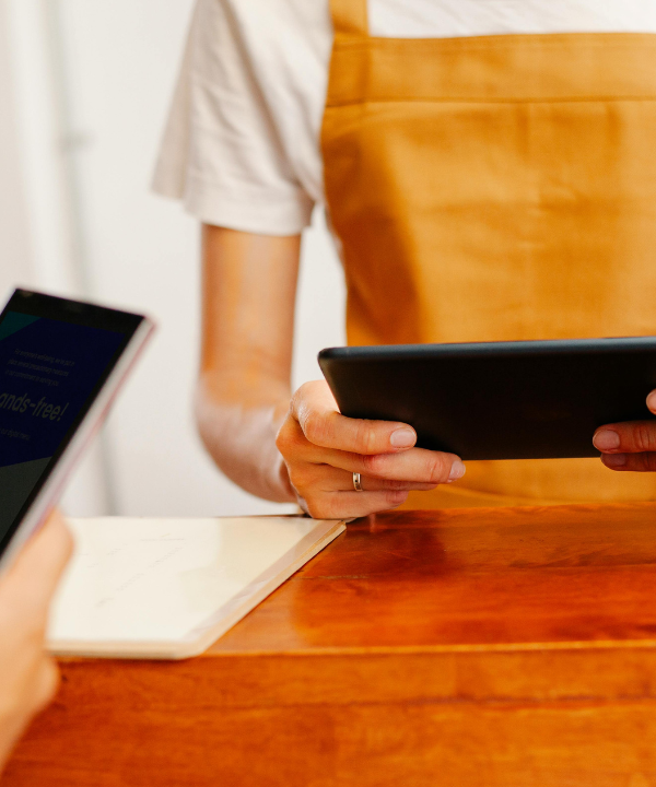 A person wearing a white t-shirt and orange apron holds an ipad while standing at a desk