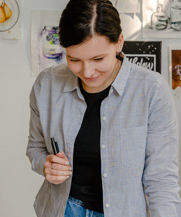 Young woman with brown hair looks down at worktable in office while holding a pen.