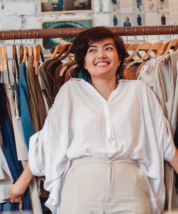 Smiling woman wearing white shirt stands in front of clothing rack in small business.