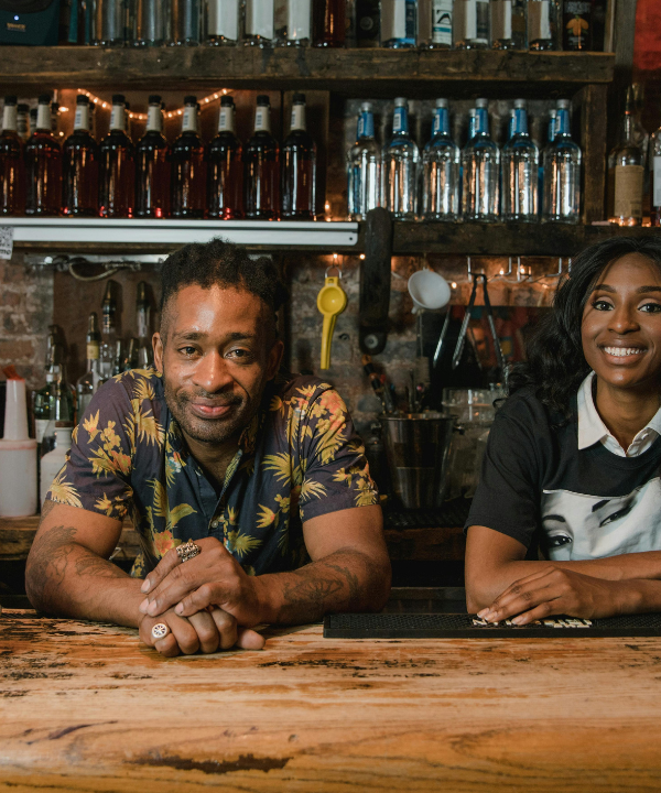 Man and woman lean on wooden bar top next to plate of food in front of bar shelving on exposed brick wall.