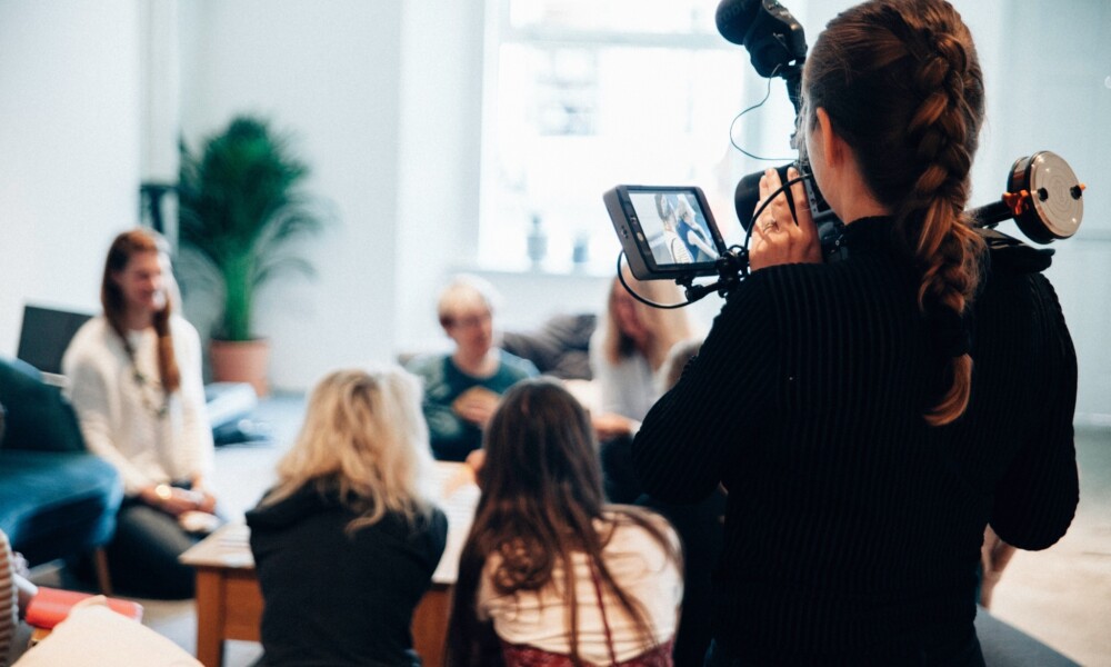 A woman holding a large camera films a group of people sitting around a table