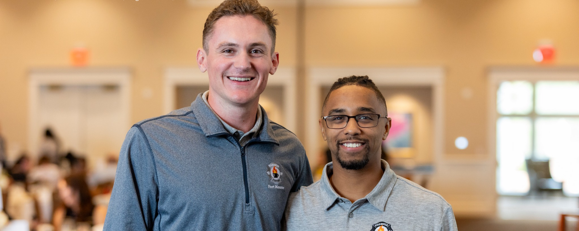 Two young men smile with backdrop of conference room with people sitting at tables.