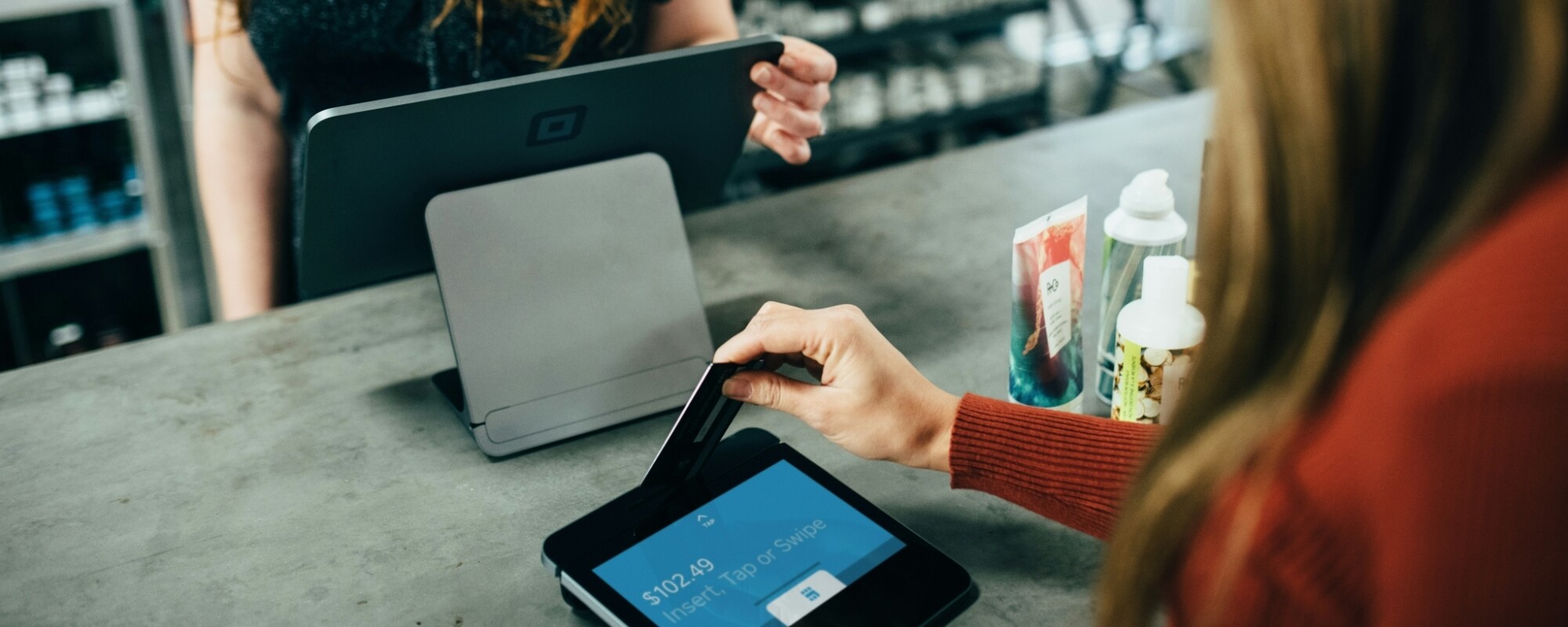 A woman stands at a cash register while another woman inserts her credit card into a payment machine