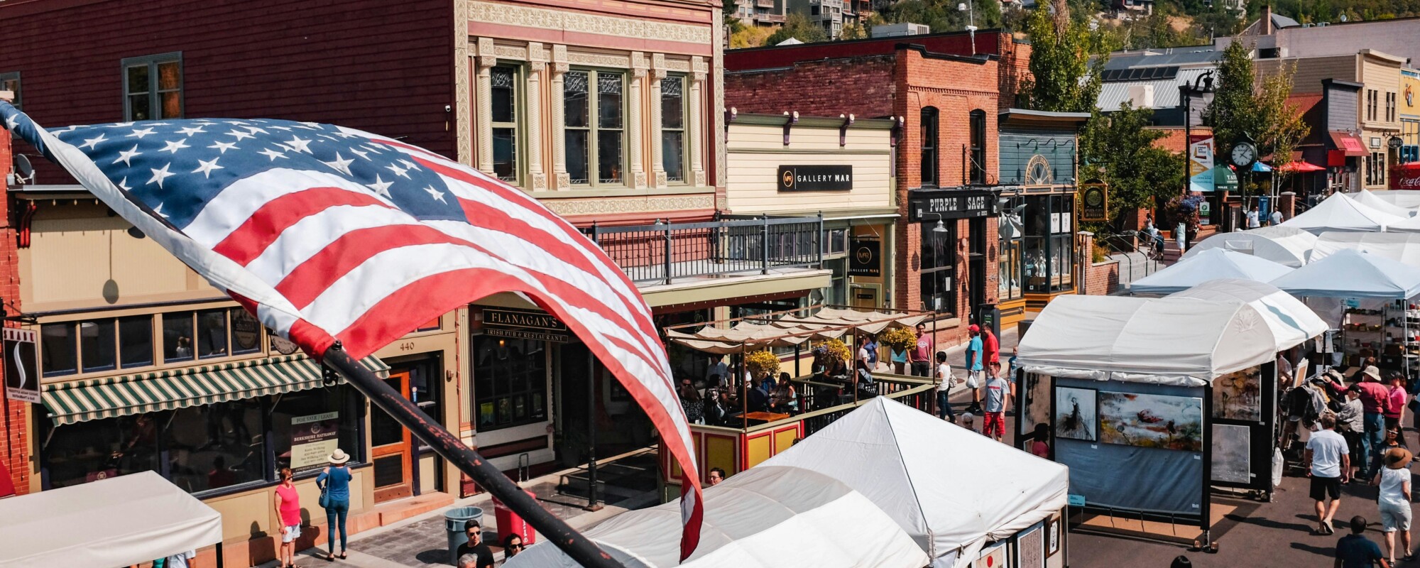 Historic Main Street during a street fair with vendors and people walking around; a large American flag waves in the foreground