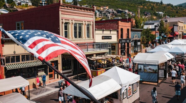 Historic Main Street during a street fair with vendors and people walking around; a large American flag waves in the foreground