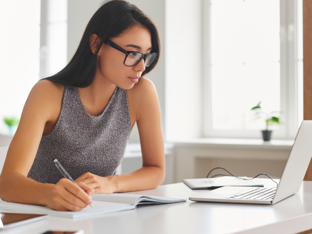 A young woman takes notes while studying at a laptop.