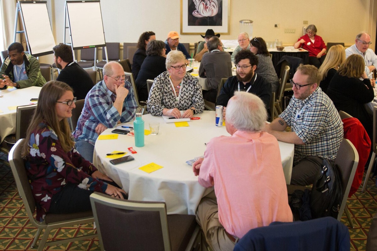 Six people sit around a table discussing course materials. In the background, other participants engage in small group discussions.