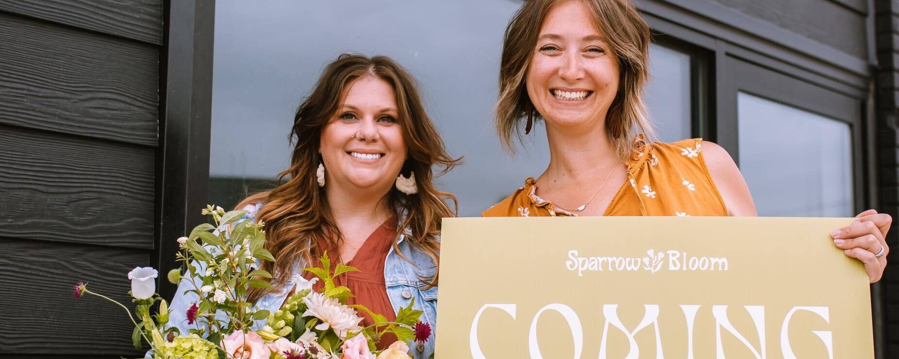 Two women pose in front of their new retail storefront: one holds a bouquet of flowers while the other holds a sign reading "Opening Soon!"