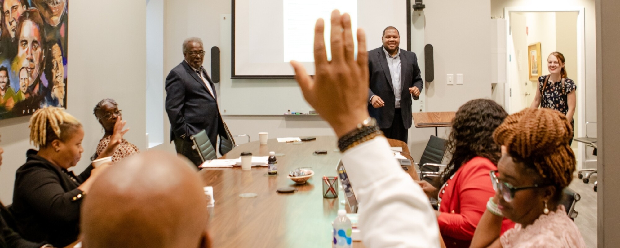 People gather around a conference table while speakers present at the front of the room.