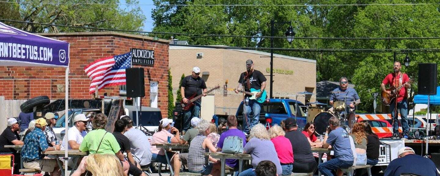 People sit at tables set-up on a street and chat while listening to a band performing on a stage.