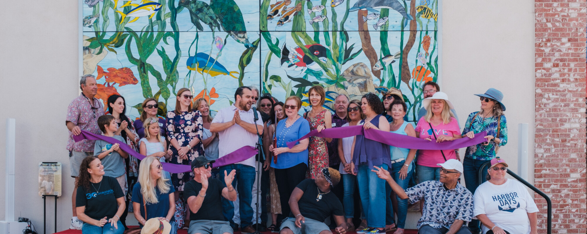 A large group of adults and children gather on a stage for a ribbon cutting event celebrating the completion of a nautical-themed mosaic mural that is visible behind them.