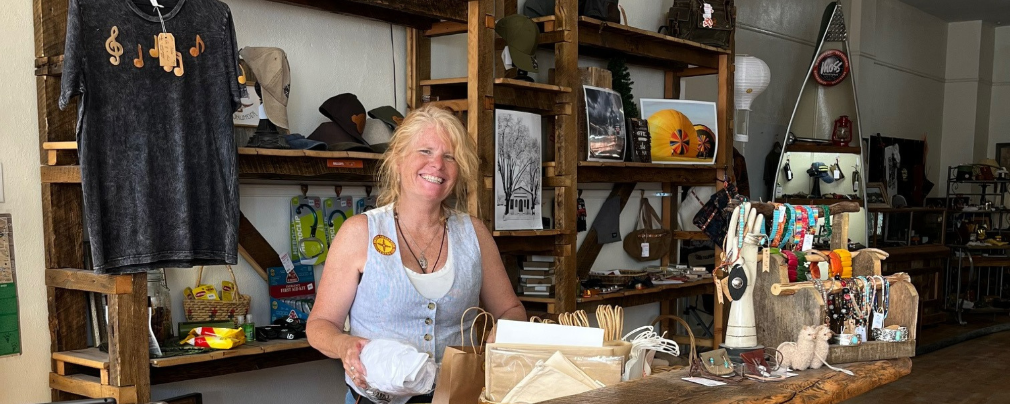 A woman smiles while standing behind a retail store counter and placing an item into a paper bag.