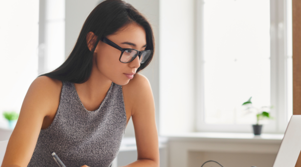 A young woman takes notes while studying at a laptop.