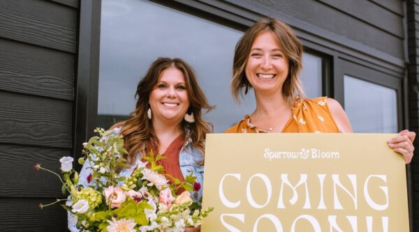 Dos mujeres posan delante de su nuevo escaparate: una sostiene un ramo de flores mientras la otra sostiene un cartel en el que se lee "¡Próxima apertura!".