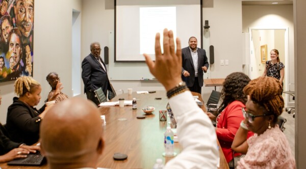 People gather around a conference table while speakers present at the front of the room.