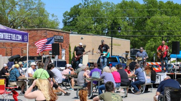 People sit at tables set-up on a street and chat while listening to a band performing on a stage.