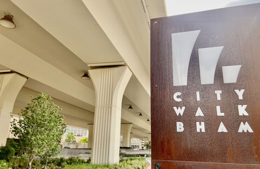 Lush pedestrian promenade beneath an overpass with a rust-colored sign reading "CITY WALK BHAM" in the foreground.