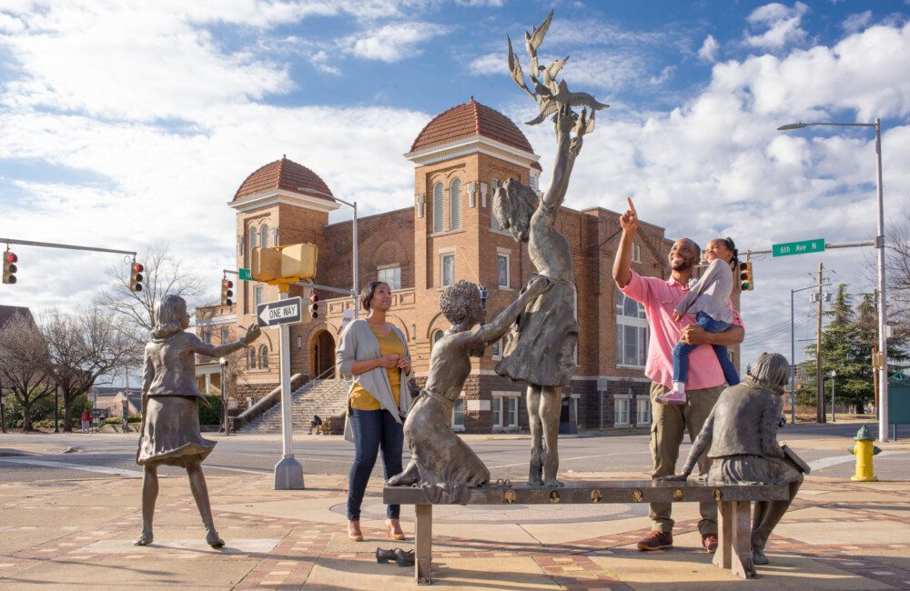 A family engages with a sculpture installation made up of four girls positioned in various poses; a large church is visible in the background.
