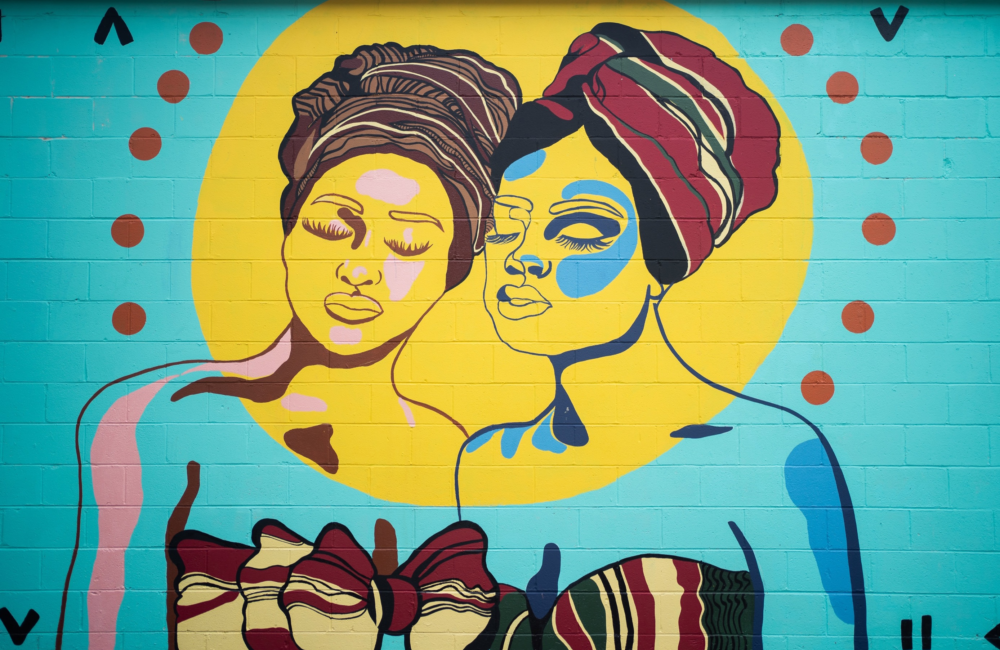 A mural painted in vibrant teal, yellow, red, and green depicting two women leaning into each other; they wear head wraps and dresses evocative of African prints.