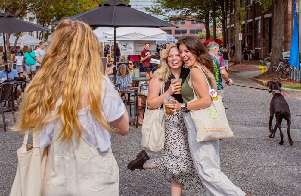 A woman takes a photo of two young women holding beverages and while embracing at an outdoor farmers market.
