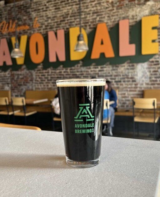 A pint of dark beer sits on a countertop; in the background "Welcome to Avondale" is painted on a brick wall in bright, bold font.