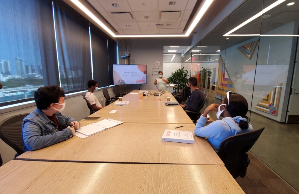 Four people sit around a conference table intently listening to a woman who is presenting from the front of the room.