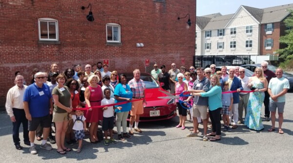 Group of people gather at official ribbon cutting ceremony in front of electric car charging station.