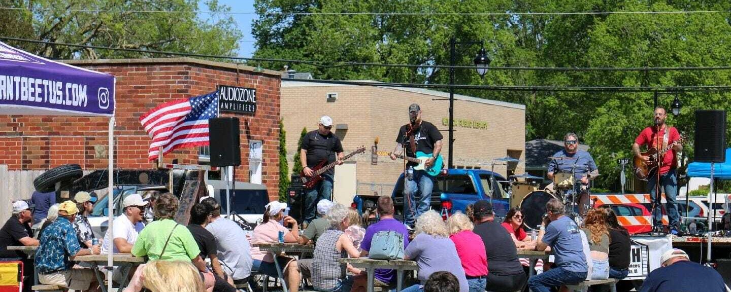 People sit at tables set-up on a street and chat while listening to a band performing on a stage.