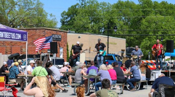 People sit at tables set-up on a street and chat while listening to a band performing on a stage.
