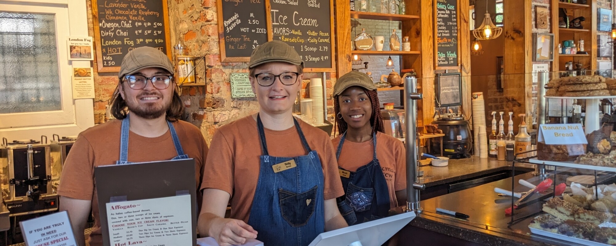 Three baristas smile while standing behind the counter of a coffee shop.