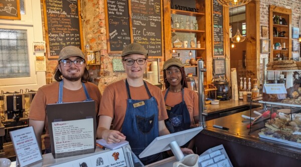 Three baristas smile while standing behind the counter of a coffee shop.