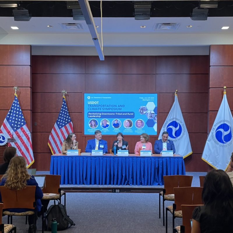 A panel of speakers sits at a table surrounded by US and DOT flags