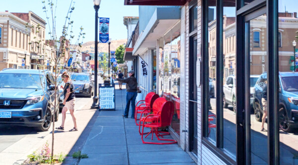 People walk along a sidewalk and a woman standing near a sign promoting a coffee business waves; bright red chairs offer seating along the exterior facade of the building.