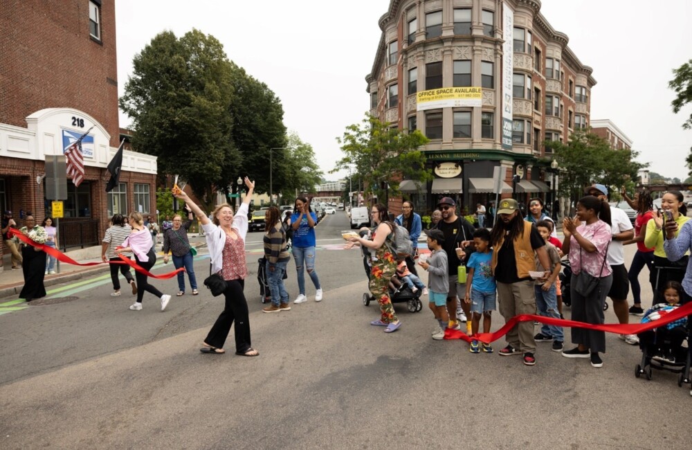 A woman raises her arms in the air after cutting the ribbon to unveil the new mural. Behind her, a crowd claps and celebrates.