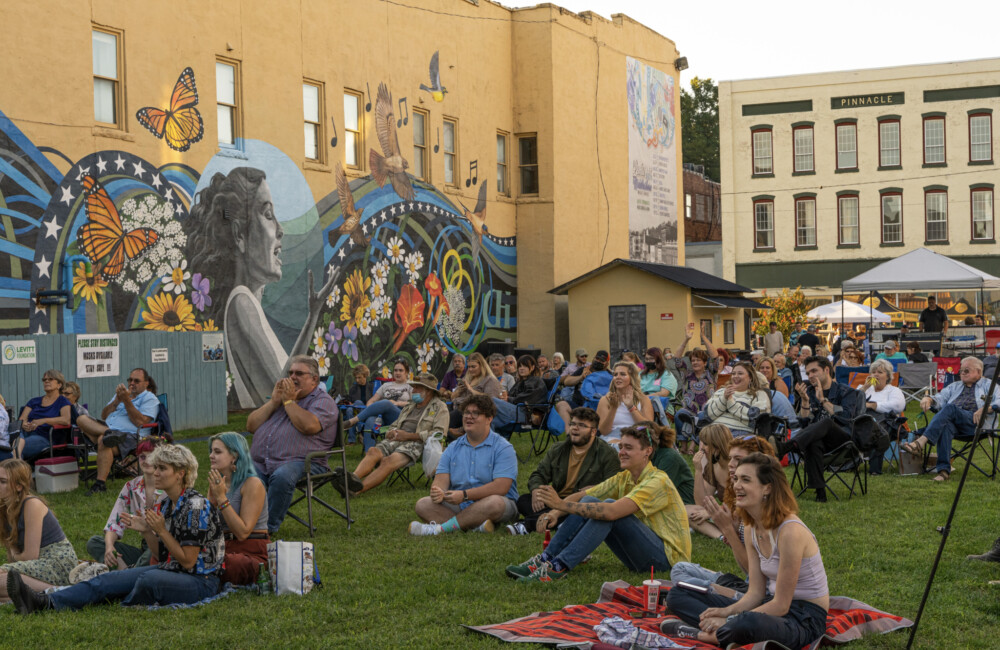 People sit on grassy lawn listening to live music in front of mural on exterior or historic building