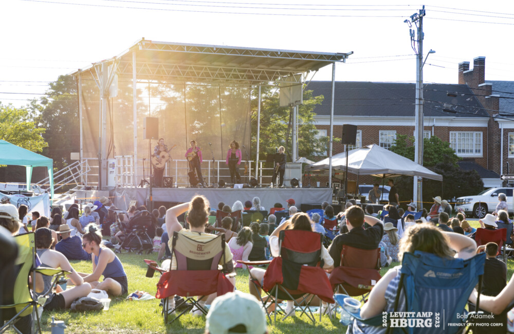 Group of people sit on grassy lawn listening to live music on outdoor stage.