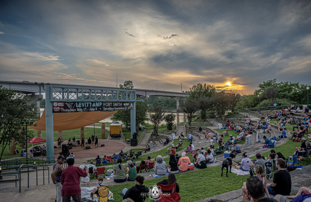 Wide angle photo of outdoor concert showing dozens of people sitting on lawn and band playing on stage.