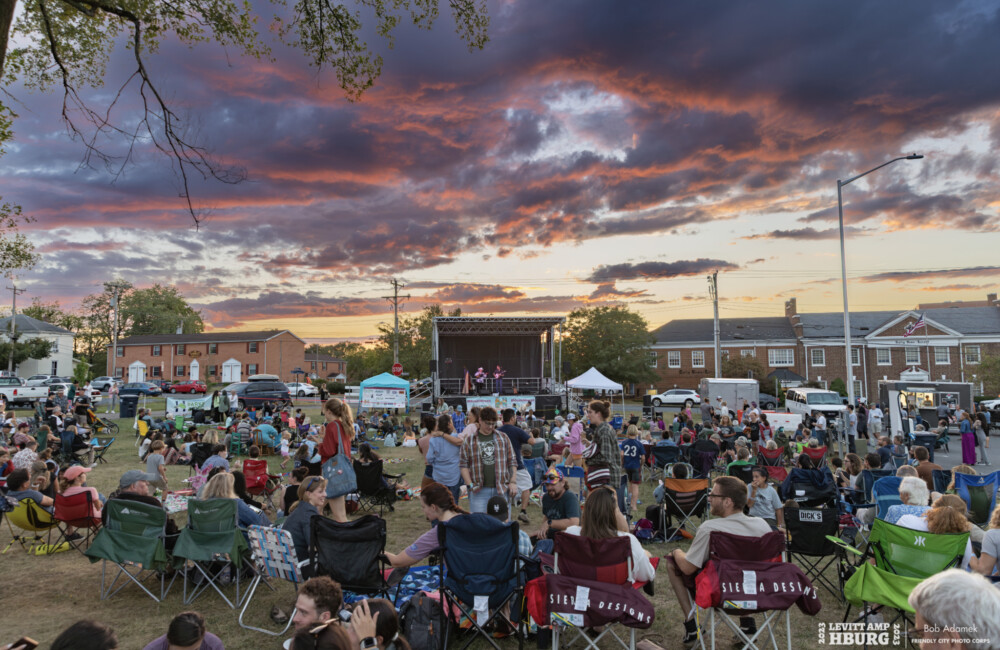 Large group of people sit on grassy lawn listening to music on outdoor stage at sunset.