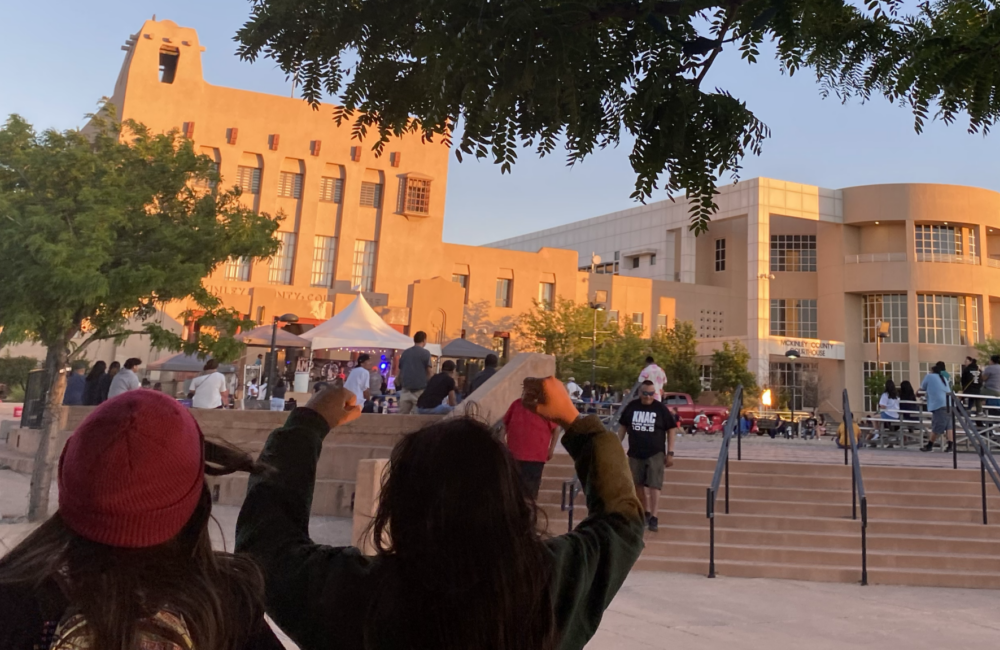 Backs of two young people cheering in foreground, group of people gathering on outdoor stage in background.