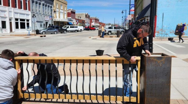 Three people working together to install a planter and fence, while in the background another person carries a square planter and main street lined with historic buildings are visible.